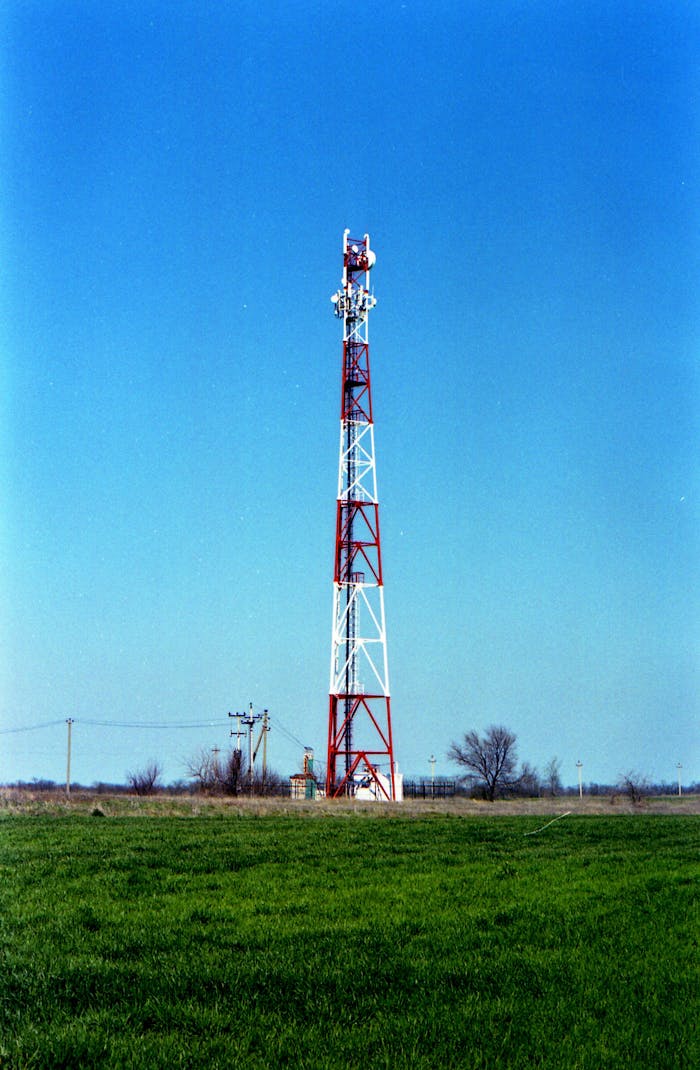 services-03 A red and white communication tower stands in a grassy field under a clear blue sky.